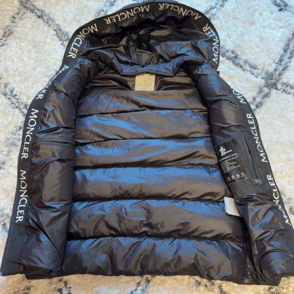 Snygg svart dunväst från Moncler med avtagbar huva och dragkedja framtill. Västen har två fickor med dragkedja och Moncler-logga på bröstet. Huva och insida har tydlig Moncler-tryck längs kanterna. Perfekt för lager-på-lager-stil.. Takit.