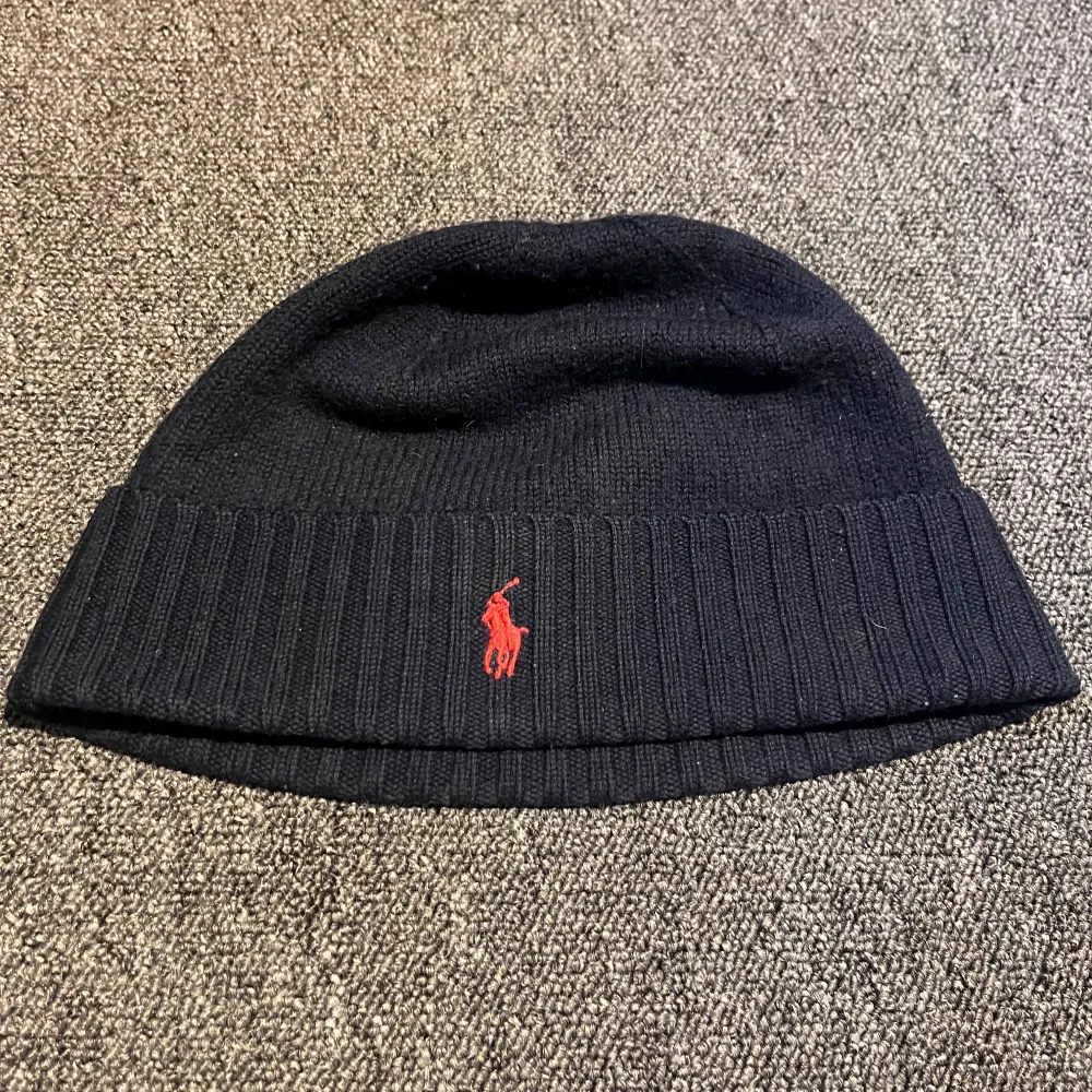 Svart stickad mössa från Polo Ralph Lauren med röd broderad logotyp framtill. Klassisk design med uppvikt kant och mjukt material som håller dig varm under kyliga dagar.. Asusteet.