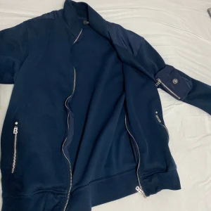 Mörkblå bomberjacka med dragkedjor - Snygg mörkblå bomberjacka med silverfärgade dragkedjor framtill och på fickorna. Jackan har lång ärm och en klassisk bomberkrage. Perfekt för dig som vill ha en stilren och enkel look.