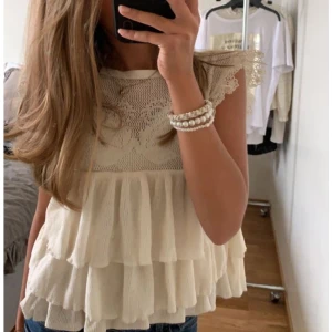 Beige volangtopp med spetsdetaljer från Zara - Supersöt beige topp från Zara med volanger och spetsdetaljer vid axlarna. Toppen har ett romantiskt mönster upptill och är ärmlös med flera lager volanger nedtill. Perfekt för en somrig look!