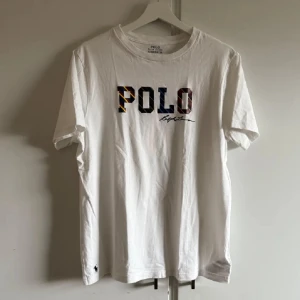 Vit t-shirt från Polo Ralph Lauren - Vit t-shirt från Polo Ralph Lauren med färgglad POLO-logga och signatur på bröstet. Klassisk passform och korta ärmar. Liten broderad logga nertill. Perfekt för en stilren och avslappnad look. Nästan aldrig använd