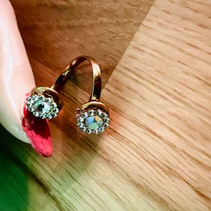 By prisförslag- Guldfärgad ring med dubbla stenar - Trendig guldfärgad ring med två runda stenar, omgivna av små gnistrande stenar. Ringen har en öppen design och passar perfekt för att ge en extra touch till din stil.