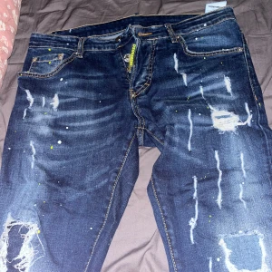 Mörkblå distressed jeans från Dsquared2 - Säljer ett par mörkblå jeans från Dsquared2 med slitningar, färgstänk och trasiga detaljer. Jeansen har klassisk femficksdesign, knappar i gylfen och tydliga kontrastsömmar. Perfekta för dig som gillar en edgy streetstil. kan gå ner på priset.