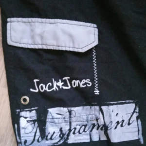 Svarta shorts Jack & Jones - Svarta shorts från Jack & Jones med gråa detaljer och grafiskt tryck i vitt och grått. Shortsen har snörning i midjan och en ficka med grå lock. Perfekta för sommaren.