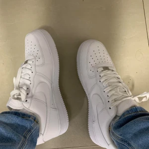 Nike Air Force 1 vita - Klassiska vita Nike Air Force 1 sneakers med låg profil och rund tå. Skorna har vit ovandel i skinn, perforerade detaljer på tån och vit sula. Perfekta för en clean och stilren look. Levereras i originalkartong.