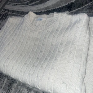 Vit kabelstickad tröja - Säljer en klassisk vit kabelstickad tröja med rund halsringning. Tröjan har lång ärm och ribbade muddar vid ärmslut och nederkant. Perfekt för lager-på-lager och passar till det mesta.