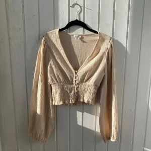 Beige blus från Monki - Säljer en snygg beige blus från Monki med v-ringning och knappar framtill. Blusen har långa ärmar och en croppad passform med elastisk midja för en bekväm stil. Perfekt för en trendig look!