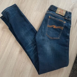 Mörkblå jeans från Nudie Jeans - Säljer ett par mörkblå jeans från Nudie Jeans med klassisk femficksdesign och orange söm på bakfickan. Jeansen har en smal passform och är tillverkade i denim med snygg tvätt. Perfekta till vardags!