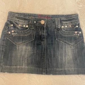 Jeanskjol med nitar och strassdetaljer - Cool jeanskjol i tvättad blå denim med snygga nitar och strass på fickorna fram och bak. Kjolen har klassisk femficksdesign, bälteshällor och knappstängning. Perfekt för dig som gillar en edgy och trendig stil.