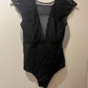 Svart spetsbody med djup v-ringning - Säljer en svart body med spetsdetaljer och djup v-ringning både fram och bak. Bodyn har ärmlös design med spets längs axlarna och meshpartier upptill. Perfekt fest topp som lätt kan kläs upp och ner. Vet tyvärr inte storleken men den passar mig som är Xs-S.  