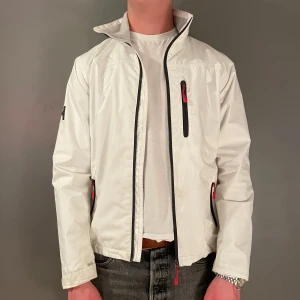Vit seglarjacka från Helly Hansen - Snygg vit vindjacka från Helly Hansen med svarta detaljer och dragkedjor. Jackan har hög krage, tre fickor med dragkedja och meshfoder på insidan. Perfekt för blåsiga dagar och enkel att matcha med olika outfits.
