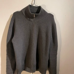 Grå stickad kofta från Maison Margiela - Säljer en grå stickad kofta från Maison Margiela med hel dragkedja framtill och hög krage. Koftan har långärmad design och ribbad struktur som ger en stilren och modern look. Perfekt för lager-på-lager under kyliga dagar.