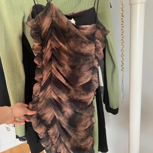 Brun och svart omlottklänning med rynkade detaljer - Säljer en kort klänning med rynkade volanger i bruna och svarta toner. Smala axelband som korsas och en tight passform. Oanvänd✨