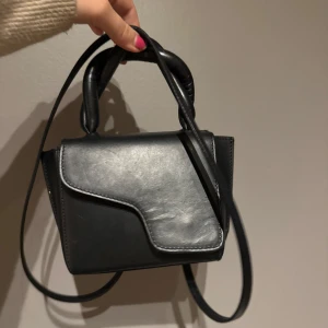 Svart handväska med flätat handtag ATP  - Snygg svart handväska med ett unikt flätat handtag och en justerbar axelrem. Väskan har en stilren design med ett lock som stängs över framsidan. Perfekt för att bära dina nödvändigheter med stil. Bra skick förutom lite stilten framtill som ni ser på andra bilden💕