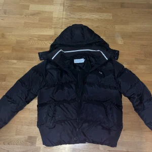 Svart pufferjacka från Calvin Klein - Säljer en svart pufferjacka från Calvin Klein med huva och dragkedja. Jackan har en normal passform och är perfekt för kyligare dagar. Klassisk design med diskret logga på bröstet och två sidofickor.