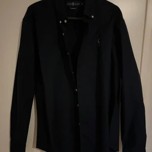 Svart skjorta från Ralph Lauren - Klassisk svart skjorta från Ralph Lauren med knappar framtill och broderad logga på bröstet. Skjortan har lång ärm och normal passform, perfekt till jeans eller chinos.