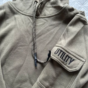 Grön hoodie med fickdetalj - Säljer en grön hoodie med praktisk ficka på ärmen märkt 'UTILITY'. Den har en justerbar dragsko i huvan och är perfekt för en avslappnad stil. Första bilden är en bättre bild på hur färgen faktiskt ser ut då det var kontogt ljus i andra bilden 🤣