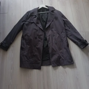 Svart trenchcoat från Batistini - Snygg svart trenchcoat från Batistini med dubbelknäppning och klassisk krage. Jackan har långa ärmar och justerbara remmar vid ärmsluten för en stilren look. Perfekt för höst och vår!