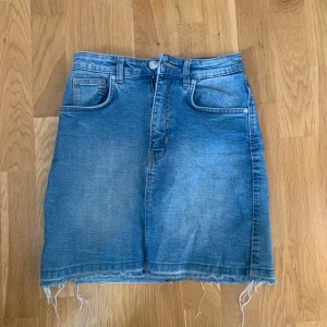 Jeanskjol  - Snygg jeanskjol i klassisk blå denim med rå kant nertill. Kjolen har femficksdesign och stängs med knapp och dragkedja framtill. Köparen står för frakten 