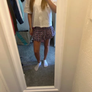 Mönstrad kortkjol i blått och rosa - Säljer en söt kortkjol med ett färgglatt mönster i blått och rosa. Kjolen har en volangdetalj som ger den en lekfull touch. Perfekt för sommarens varma dagar!