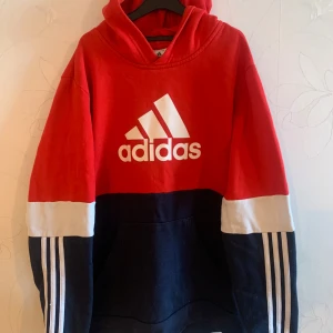 Röd hoodie från Adidas - Säljer en snygg röd och marinblå hoodie från Adidas med det klassiska logotyptrycket på framsidan. Tröjan har långa ärmar med vita ränder längs sidorna och en bekväm huva. Perfekt för en sportig look! Priset går såklart att diskutera!