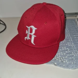 Röd keps med broderad bokstav - Snygg röd keps med en vit broderad bokstav på framsidan. Perfekt för att ge din outfit en cool touch. Justerbar baksida för bästa passform.