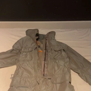 Grå vindjacka från Parajumper - En grå windbreaker från Parajumper med flera fickor och dragkedjor. Jackan har en hög krage och justerbar midja. Perfekt för blåsiga dagar. //Trendz.resell!
