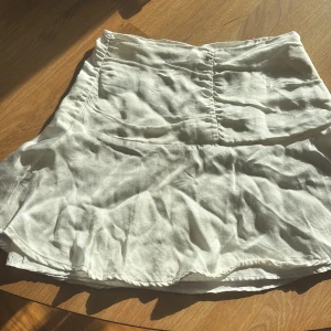 Vit silk kortkjol från Bershka - Säljer en vit kortkjol från Bershka i lätt och mjukt material. Kjolen har en diskret dragkedja i sidan och en lätt utsvängd form som ger en avslappnad look. Perfekt för varma dagar och enkel att matcha med olika toppar.