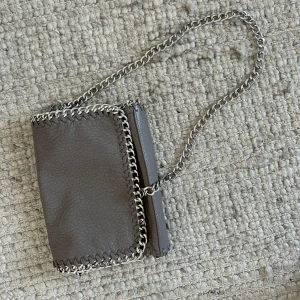 Grå axelväska med kedjedetaljer från Tiamo - Säljer en grå axelväska från Tiamo med silverfärgad kedja som axelrem och runt kanterna. Väskan är i skinnimitation och har dragkedja på baksidan samt magnetknappar för stängning. Perfekt för att lyfta din outfit!