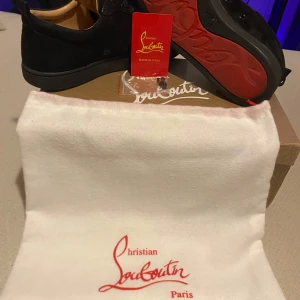 Svarta sneakers från Christian Louboutin - Snygga svarta sneakers från Christian Louboutin med ikonisk röd sula och nitar på tån. Skorna har snörning och är tillverkade i mocka. Perfekta för att ge din stil en edgy touch.