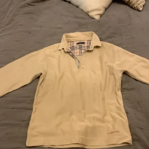 Beige pikétröja från Burberry - Säljer en stilren beige pikétröja från Burberry med klassiskt rutigt mönster på insidan av kragen. Tröjan har en dragkedja framtill och långa ärmar. Perfekt för en avslappnad men ändå elegant look. Nästan aldrig använd och passar längden 165-170