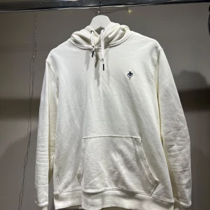 Vit hoodie från Morris - Säljer en stilren vit hoodie från Morris med en liten broderad logga på bröstet. Tröjan har en klassisk känguruficka och justerbar huva med snören. Perfekt för en avslappnad stil.