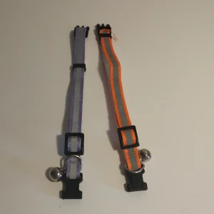 Två randiga halsband med bjällra - Säljer två justerbara halsband med bjällra, ett i lila och ett i orange. Båda har ett randigt mönster och är utrustade med ett svart plastspänne för enkel på- och avtagning. Perfekt för att hålla koll på ditt husdjur med stil.