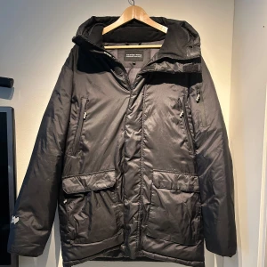 Svart dunjacka från Mountain Works - Säljer en svart dunjacka från Mountain Works. Jackan har en praktisk huva och dragkedja framtill. Den är utrustad med flera fickor för extra förvaring. Perfekt för kyliga dagar. Nypris 4000kr, priset ej hugget i sten