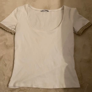 Vit topp med spetsdetaljer från Zara - Säljer en söt vit topp från Zara med korta ärmar och spetsdetaljer vid ärmsluten. Toppen har en rundad halsringning och är perfekt för en stilren look.
