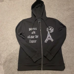 Svart hoodie från Blinglucid - Snygg svart hoodie från Blinglucid med texten 'paris au clair de lune' i strass på framsidan. Hoodien har en dragkedja och en cool design med Eiffeltornet i strass. Perfekt för dig som vill sticka ut med stil!