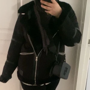 Svart jacka med pälsdetaljer - Håll vid arm, ej synlig. Snygg svart jacka från pull and bear med pälsdetaljer på kragen och dragkedjor framtill. Jackan har en cool bikerstil med en asymmetrisk dragkedja och spänne vid kragen. Perfekt för att hålla stilen i kallare väder. 