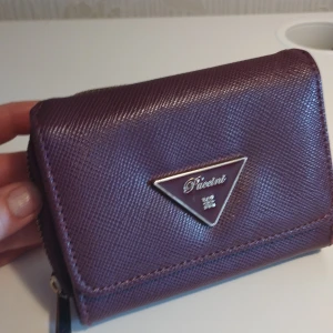 Lila plånbok från Puccini - Snygg lila plånbok från Puccini med flera fack för kort och en dragkedja för mynt. Den har en elegant triangulär logotyp på framsidan och är tillverkad i syntetmaterial med en texturerad yta. Perfekt för att hålla ordning på dina kort och kontanter.