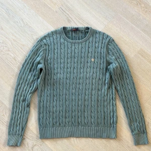 Grön stickad tröja från Morris - Säljer en stilren grön stickad tröja från Morris med kabelstickat mönster. Tröjan har långa ärmar och ribbade muddar vid ärmslut och nederkant. Perfekt för kyligare dagar!