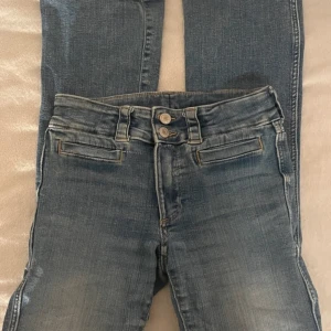Blå bootcut jeans från H&M - Snygga blå bootcut jeans från H&M med låg midja och justerbar midja. De har en klassisk femficksdesign och stängs med två knappar och dragkedja. Perfekta för en avslappnad stil.