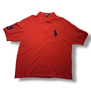 Röd pikétröja från Ralph Lauren - Säljer en snygg röd pikétröja från Ralph Lauren med klassisk krage och knappar. Tröjan har en blå logotyp på bröstet och en siffra på ärmen. 