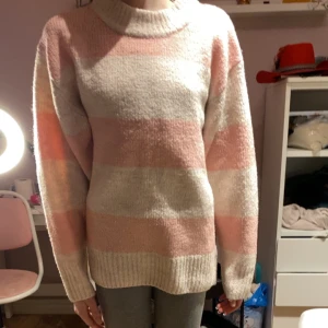 Rosa och vit randig stickad tröja - Mysig stickad tröja i rosa och vitt med breda ränder. Tröjan har en hög krage och långa ärmar, perfekt för kyligare dagar. Den är både stilren och bekväm, vilket gör den till ett utmärkt val för höst och vinter.