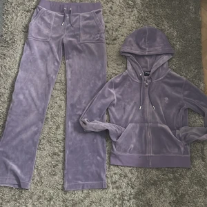 Lila juicy mjukisbyxor med matchande lila kofta  - Säljer ett set med lila juicy mjukisbyxor och matchande kofta. Byxorna har en bekväm passform och snörning i midjan samt fickor fram och bak. Koftan har dragkedja och en mysig huva. Perfekt för en avslappnad stil. Har andvändts få gånger. Har en liten defekt då en metallbit på ett av snörena på byxorna har gått av, det syns inte alls mycket. Kontakta även ifall köp av enskilt plagg. 