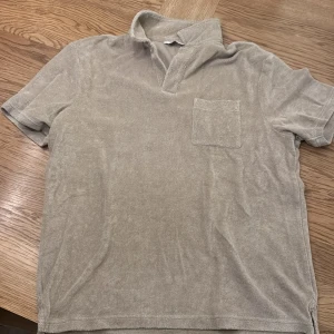 Beige frotté t-shirt från East West - Säljer en beige frotté t-shirt från East West. Den har en klassisk krage och en bröstficka. Perfekt för en avslappnad stil. Passar bra till både jeans, linnebyxor och shorts.