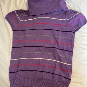 Lila randig tröja från Colours of the World - Säljer en lila tröja från Colours of the World med färgglada ränder i vitt, rött, grönt och mörklila. Tröjan har korta ärmar och en hög krage som ger en unik stil. Perfekt för att liva upp garderoben!🤍 #fintröja #kortarmad #lilatröja #randig #krage #coloursoftheworld