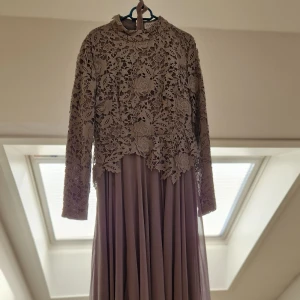 Grå aftonklänning med spetsdetaljer - Elegant grå aftonklänning med vackra spetsdetaljer och långärmad design. Perfekt för en stilfull look med en flödande kjol som ger en romantisk känsla. Klänningen har en hög hals och ett intrikat blommönster i spetsen.