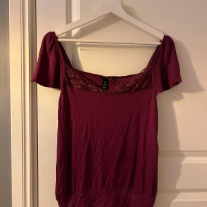 Vinröd topp med spetsdetaljer - Säljer en vinröd topp med spetsdetaljer vid halsringningen. Toppen har korta ärmar och en bekväm passform. Perfekt för att lägga till en färgklick i garderoben. Hör av dig vid intresse! 💜