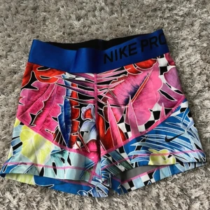 Färgglada träningsshorts från Nike - Snygga och färgglada träningsshorts från Nike med ett tropiskt blommönster i rosa, blått, gult och svart. De har en bred elastisk midja med Nike Pro-logga. Perfekta för en energisk träningssession.