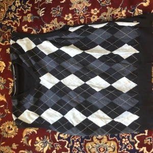 Rutig väst - Säljer en snygg rutig tröja från Wolsey i svart, grått och vitt. Tröjan har ett klassiskt argyle-mönster och är perfekt för en stilren look. Den har långa ärmar och en rund halsringning.