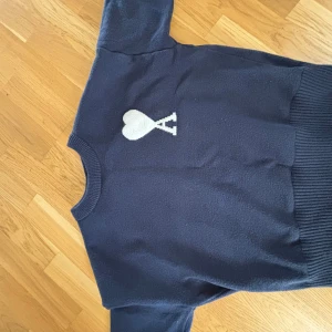 Mörkblå ami paris  tröja med hjärtdetalj - Säljer en mörkblå tröja med en vit hjärtdetalj på bröstet. Tröjan har långa ärmar och ribbade muddar vid ärmslut och nederkant. Perfekt för en stilren och bekväm look. Köpt från CN fans 